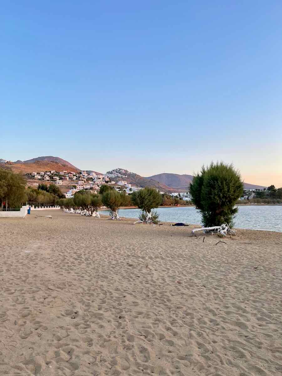 The image shows a peaceful, sandy beach with a few scattered trees lining the shore. In the background, a small coastal town with white buildings sits on the hillside under a clear blue sky. The scene captures a calm, relaxing beach atmosphere with mountains visible in the distance. It likely depicts a serene Mediterranean location.