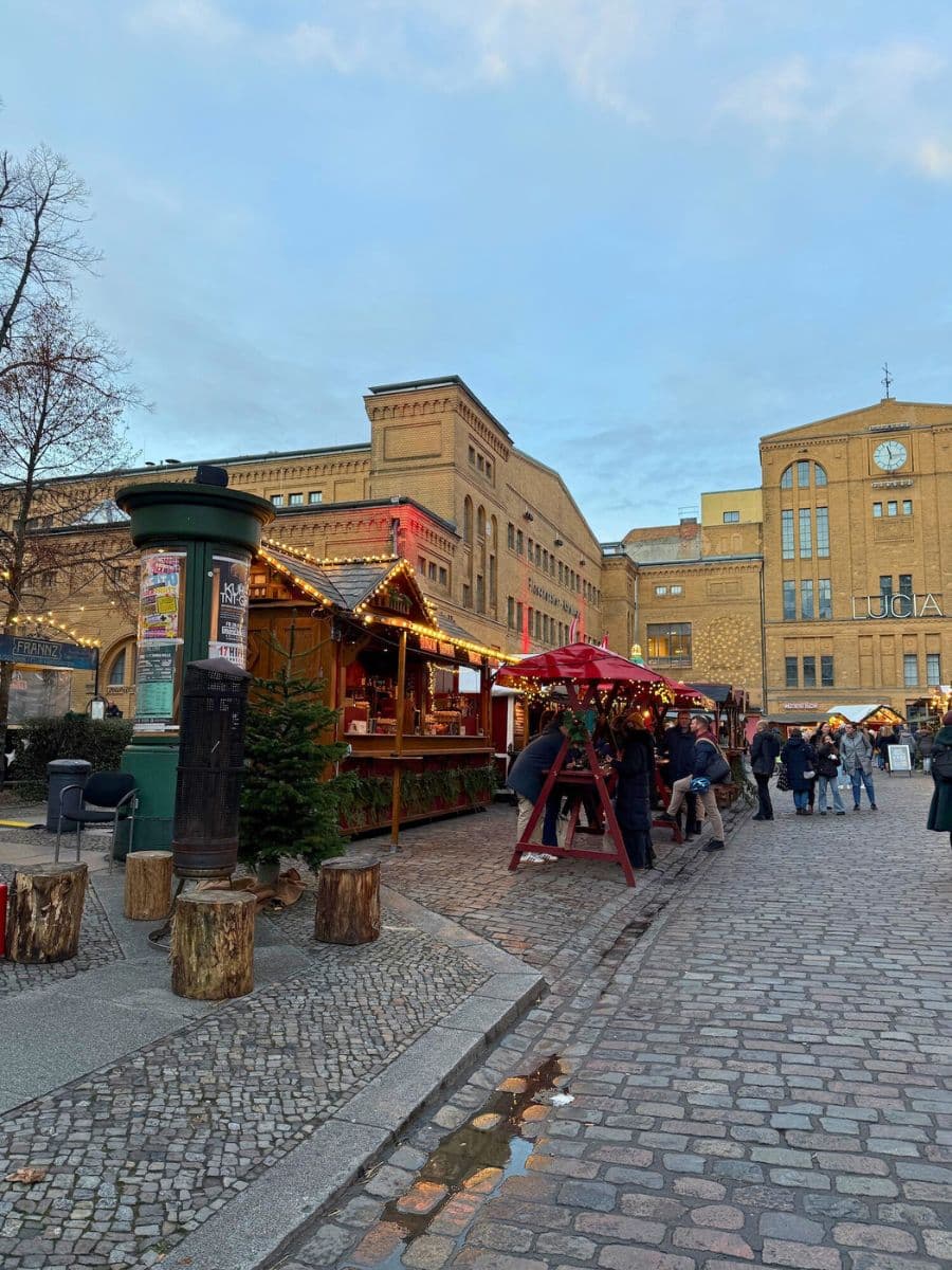 A wide view of a Berlin Christmas market with cozy wooden stalls illuminated by festive lights, surrounded by historic brick buildings and cobblestone streets.