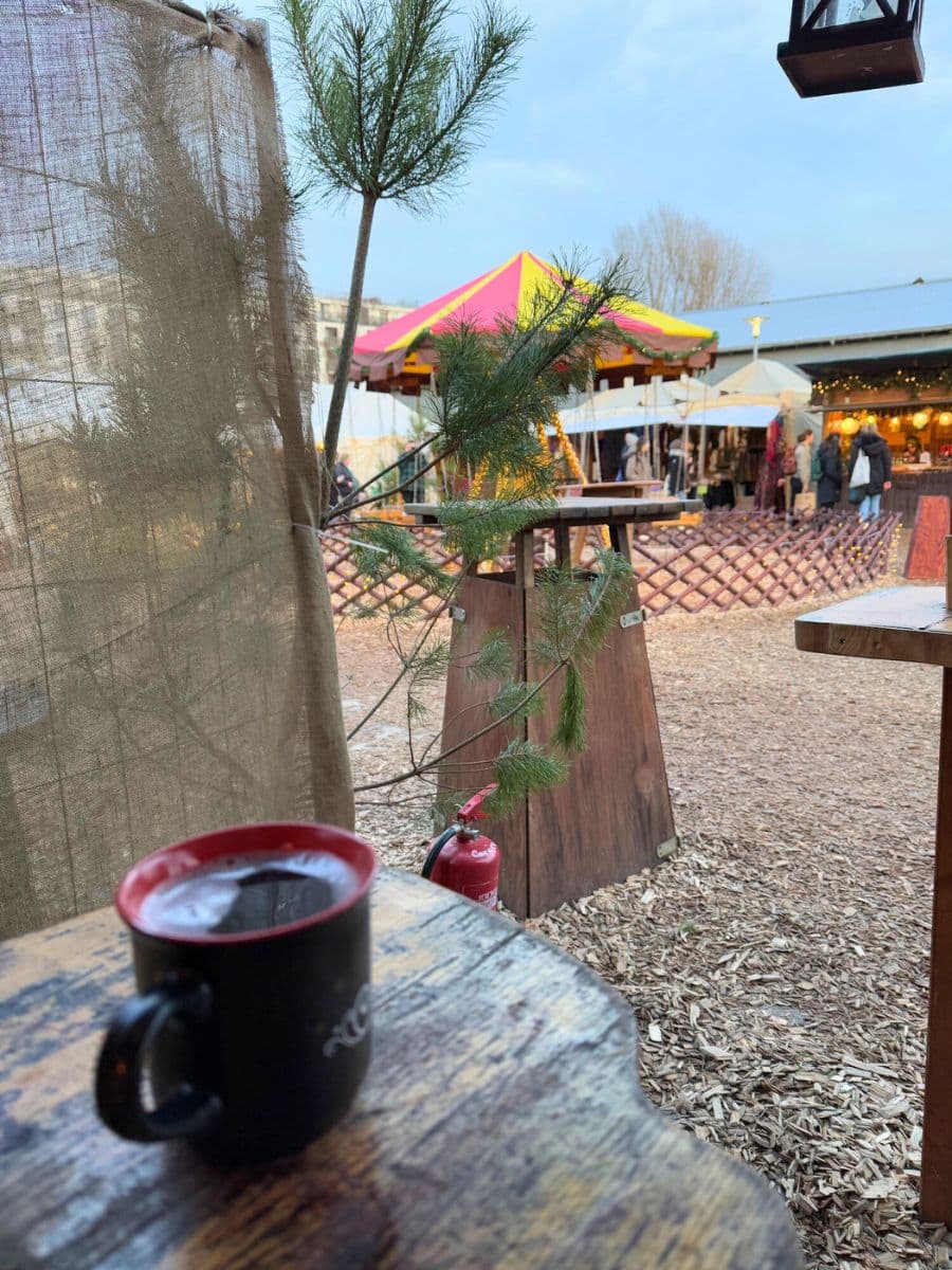 A rustic scene featuring a black mug of glühwein on a wooden table, with a colorful carousel and market tents visible in the background.