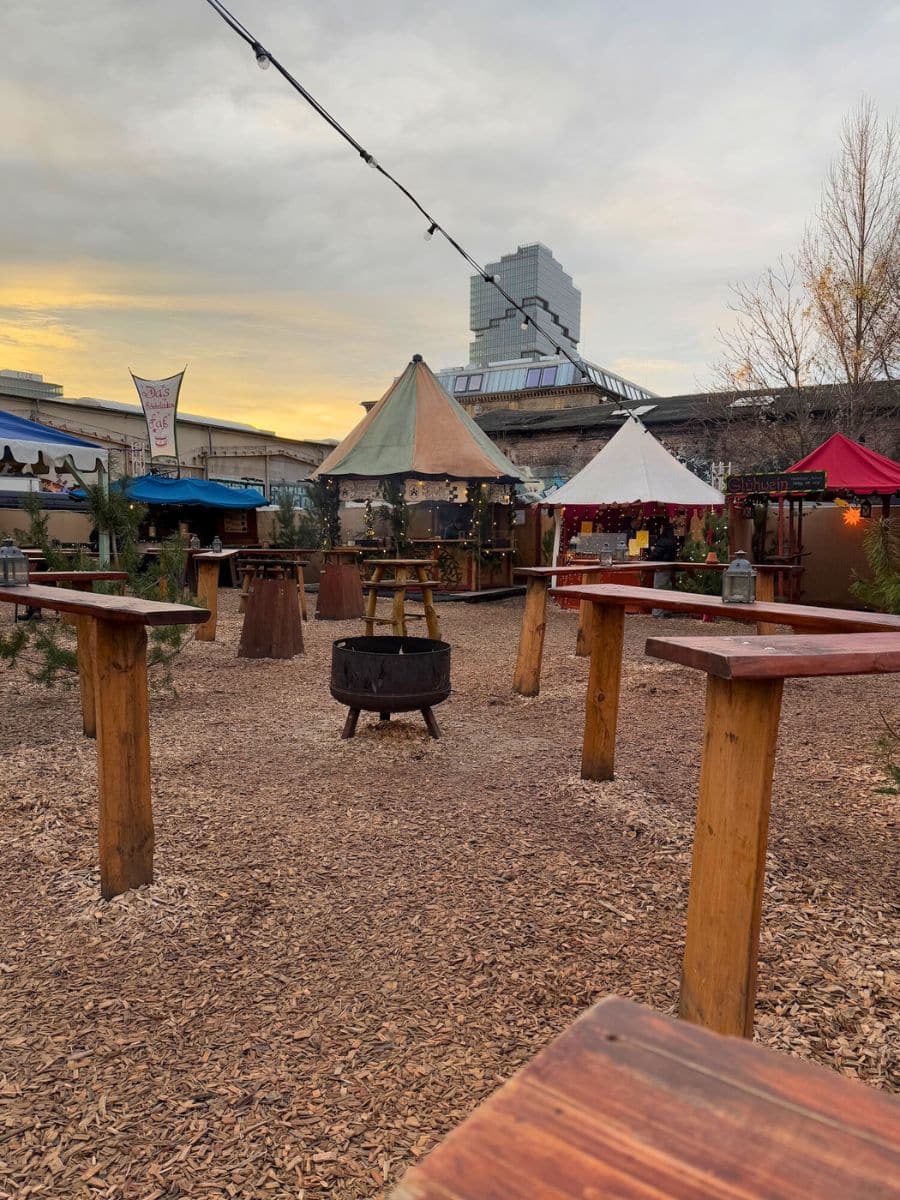 An open-air Christmas market scene with wooden tables, string lights, and colorful market tents set against a cloudy evening sky.