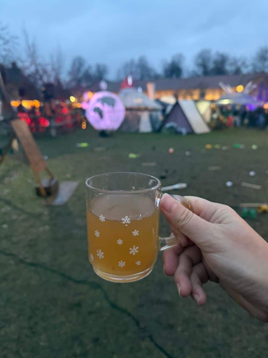 A glass mug of hot cider with snowflake patterns, held up against the backdrop of festive tents and glowing lights at the Britz Castle Medieval Christmas Market.