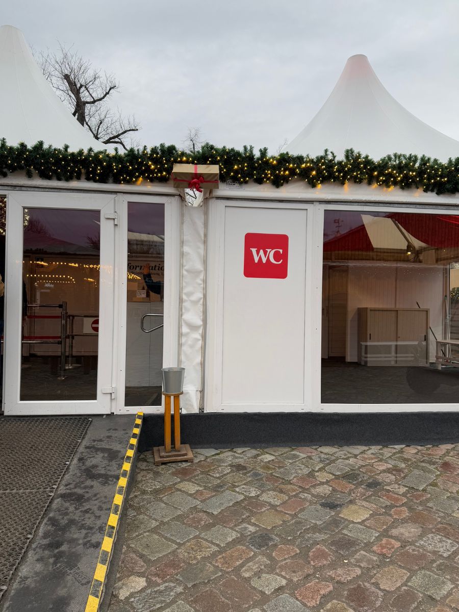 Exterior of a temporary restroom facility at a Christmas market in Berlin. White tent building decorated with garland and lights, with a red WC sign on the door.
