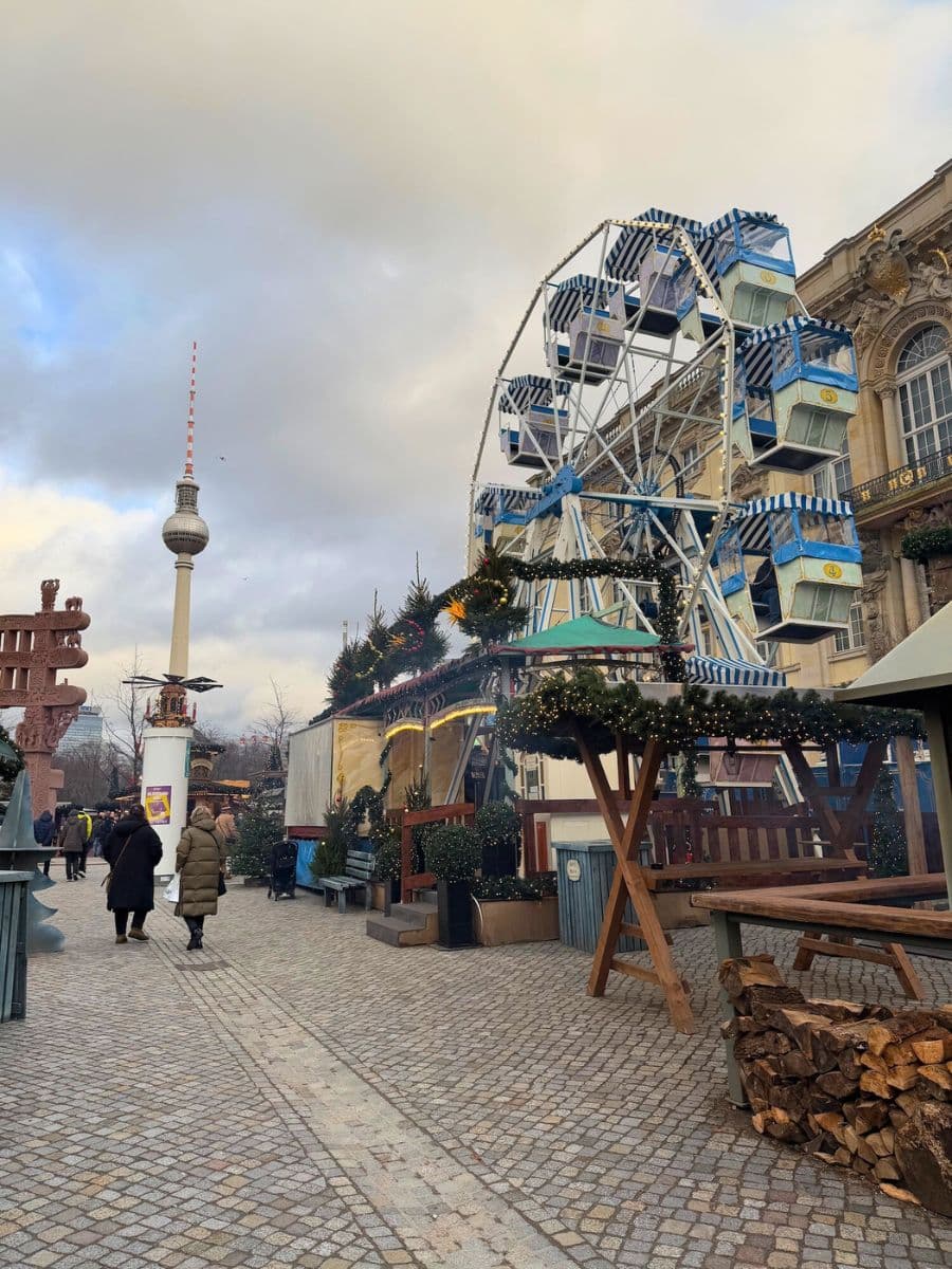 A small Ferris wheel with blue and white cabins surrounded by Christmas stalls, set against the backdrop of the Berlin TV Tower at the Humboldt Forum Christmas Market.