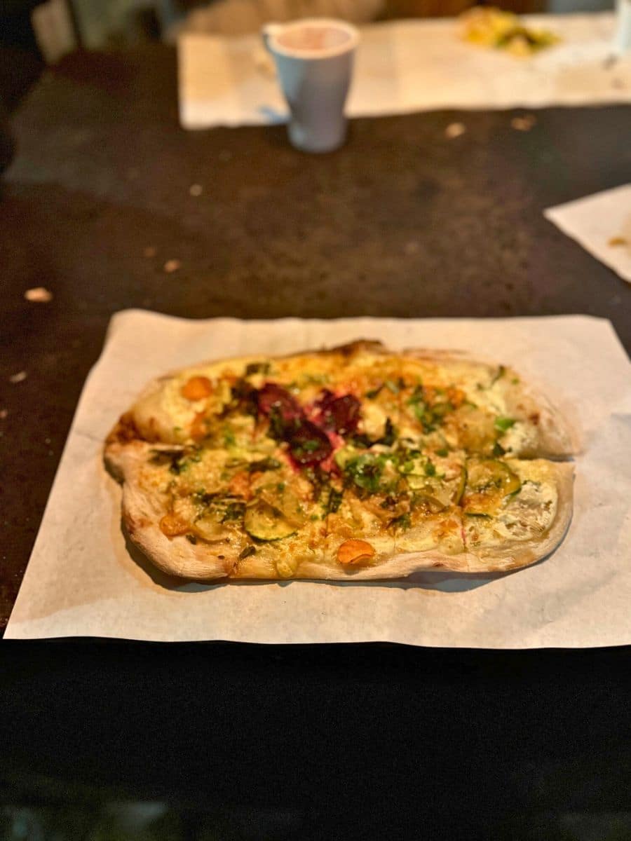 A rustic flatbread pizza served on parchment paper, with toppings including zucchini and beets, accompanied by a warm drink in a cozy market setting.