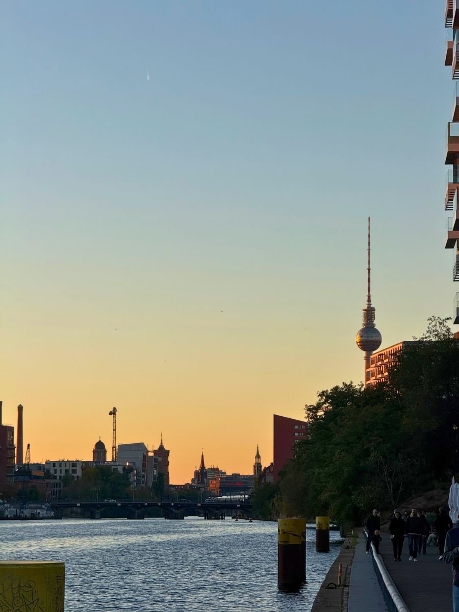 A warm, golden sunset over the Spree River in Berlin, with the TV Tower silhouetted on the horizon and pedestrians walking along the waterfront path.