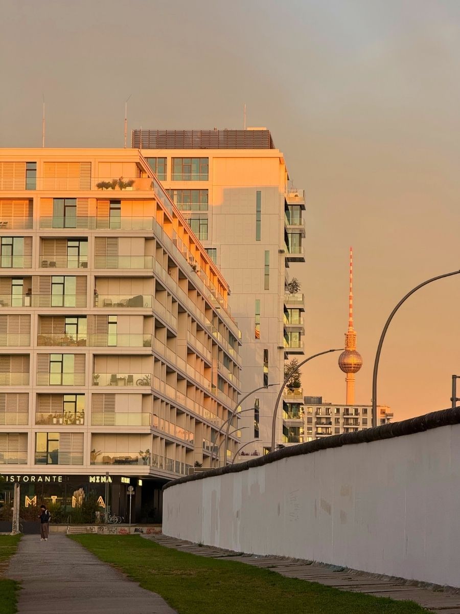 Golden hour view in Berlin showing a white modern apartment building next to a preserved section of the Berlin Wall, with the TV Tower visible in the background.