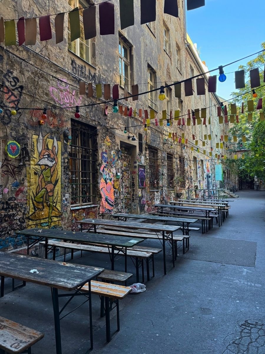 Outdoor seating area in a graffiti-covered courtyard in Berlin with long wooden tables and benches, strung with colorful prayer flags and globe lights.