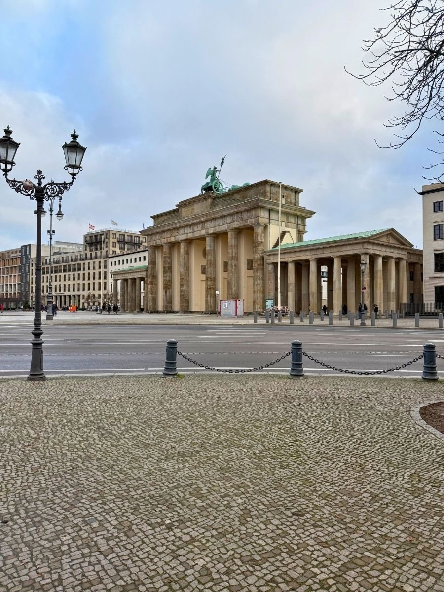 Brandenburg Gate in Berlin seen from across the plaza with cobblestone pavement and city buildings in the background.