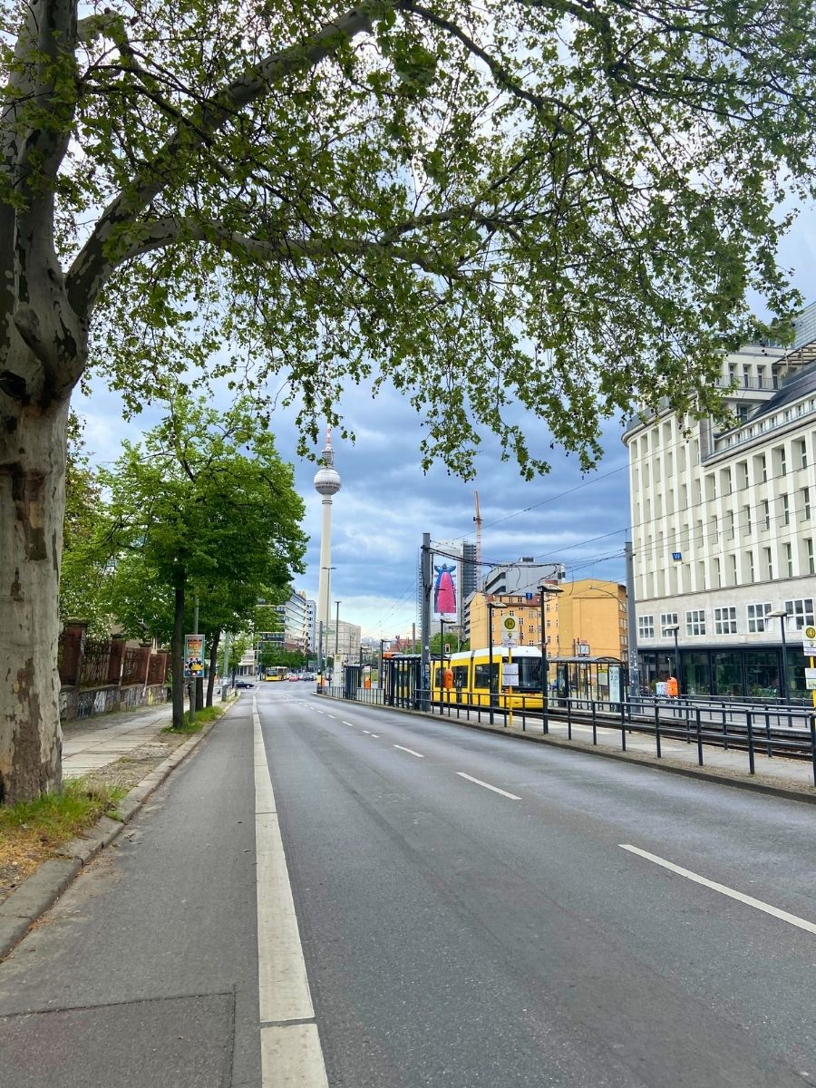 Wide Berlin street with a yellow tram and the Fernsehturm TV Tower visible in the background beneath cloudy skies.