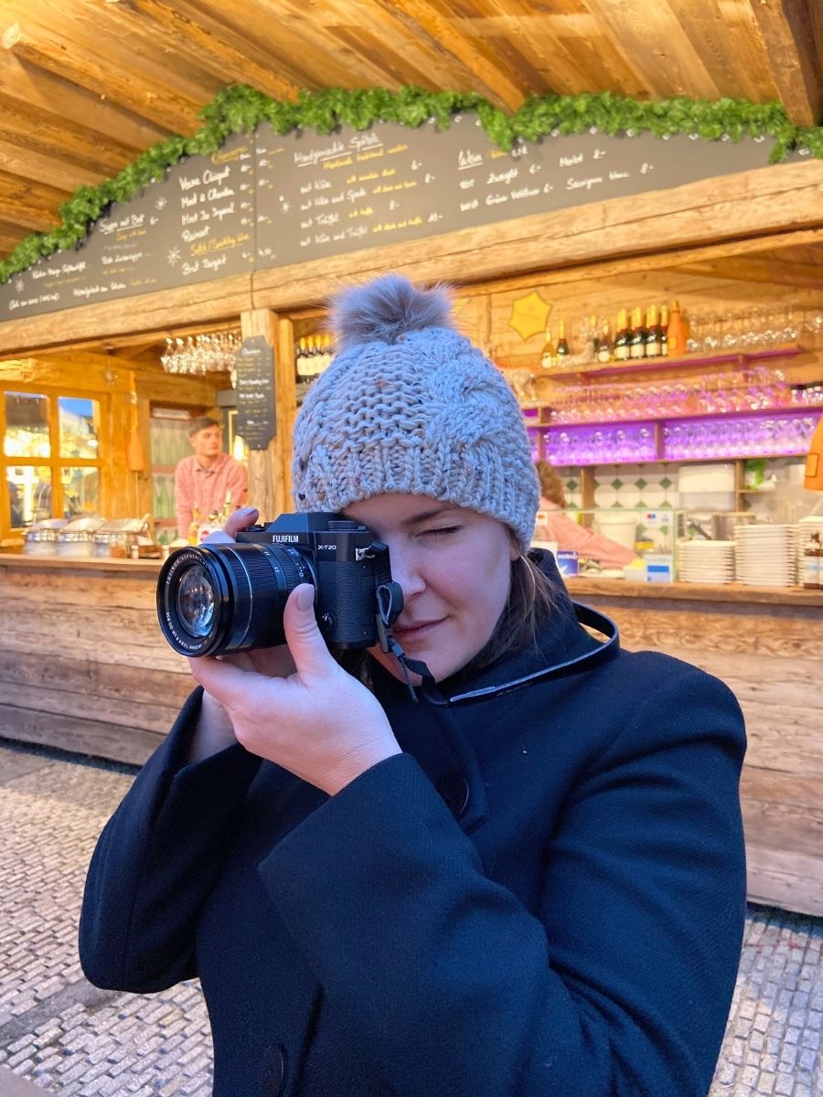 A woman in a knit hat photographing with a camera at a wooden food stall in a Berlin Christmas market.