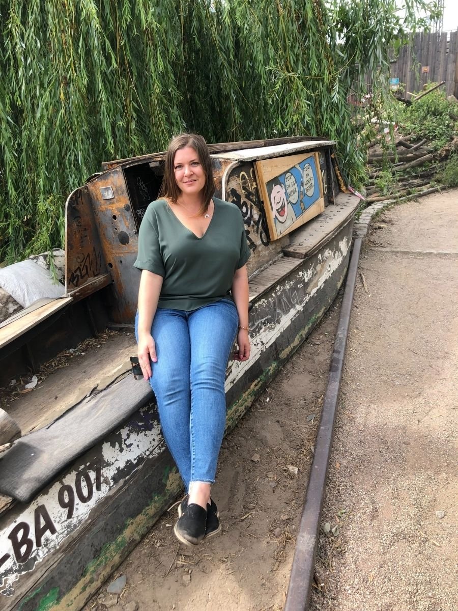 Melissa sitting on a weathered boat decorated with graffiti at an outdoor area in Berlin, surrounded by greenery.