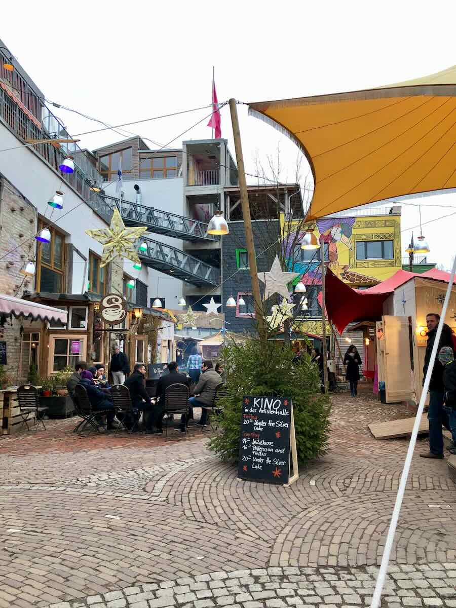 An eclectic outdoor space at the Holzmarkt in Berlin, featuring colorful signs, tents, and string lights, with people enjoying food and drinks in the cozy market atmosphere.