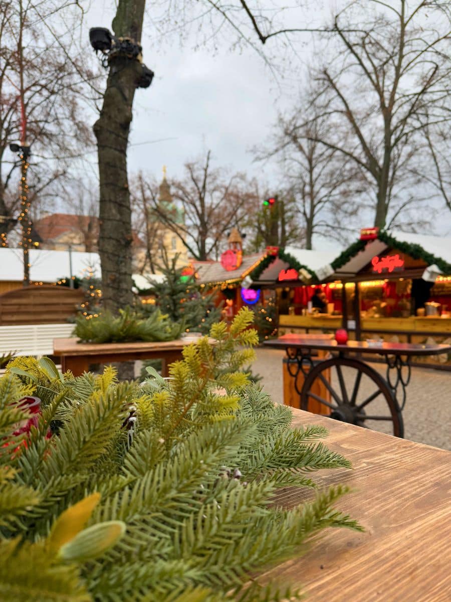 Close-up of green fir branches on a wooden table at the Christmas market. In the softly blurred background, wooden stalls decorated with lights and festive garlands are visible, along with trees and Charlottenburg Palace’s dome peeking through.