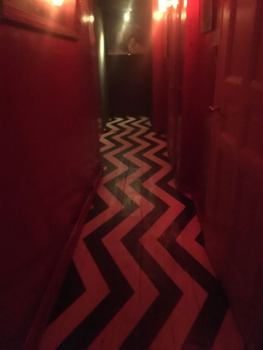 Narrow red hallway with dim lighting and a black-and-white zigzag floor pattern inspired by Twin Peaks, creating a moody, surreal atmosphere.