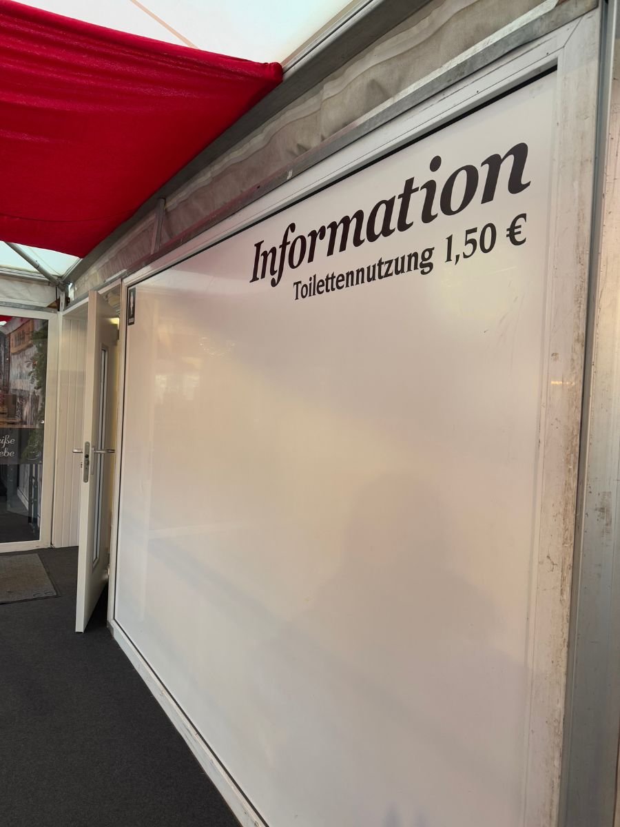 Interior wall sign at a Christmas market in Berlin showing toilet usage fee of 1.50 euros, next to the word ‘Information.