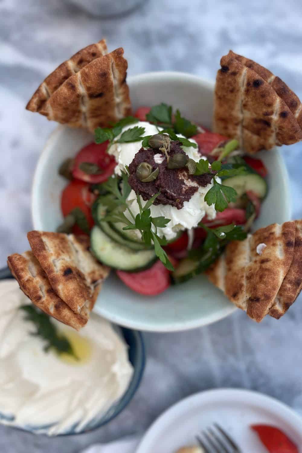 A rustic Greek salad adorned with capers and a rich dollop of tzatziki, accompanied by wedged pita bread artistically placed, served alongside a bowl of creamy spread with a drizzle of olive oil, all set on a marble table