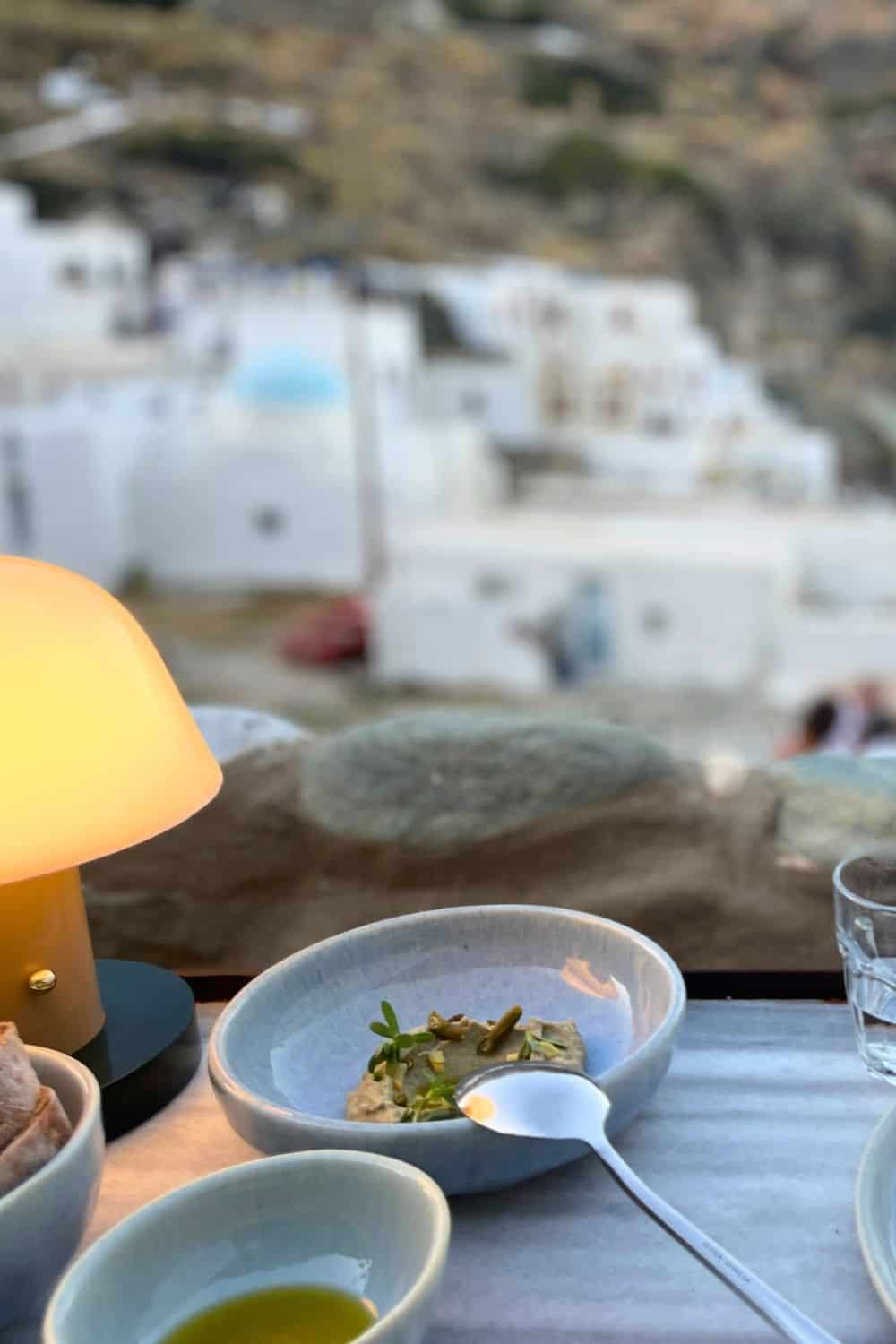 Close-up of a gourmet dish on a table with a blurry backdrop of white Greek houses in the evening light.