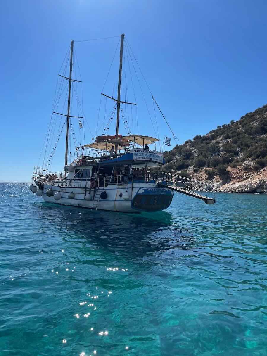 The image shows a large sailboat anchored in the crystal-clear turquoise waters off the coast of Milos, Greece. The boat has two masts and a covered deck area, with hills and rocky terrain visible in the background under a clear blue sky.