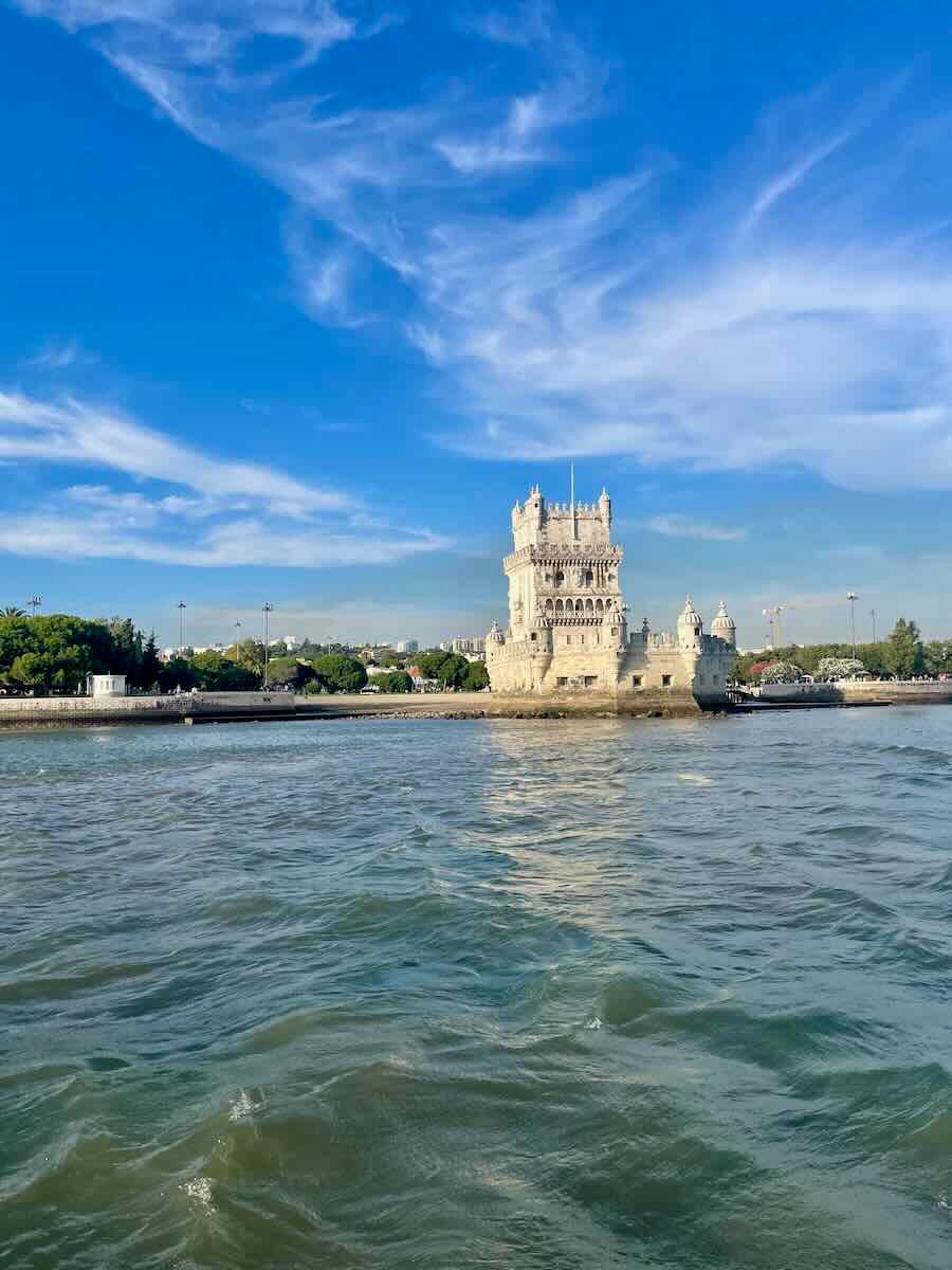 A historic stone tower, known as the Belém Tower, stands majestically on the waterfront under a bright blue sky with wispy clouds. The water below is gently rippling, reflecting the sunlight. The tower’s detailed architecture is illuminated by the afternoon light, and lush green trees can be seen in the background, along with parts of the Lisbon shoreline.