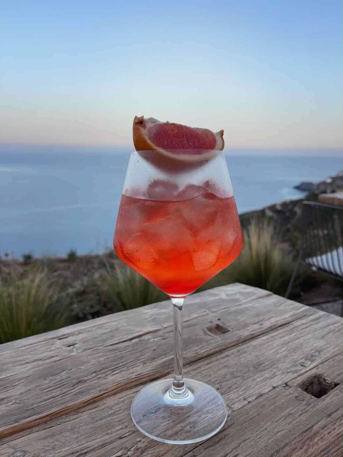 The image shows a vibrant orange cocktail served in a large wine glass with ice, garnished with a slice of grapefruit. The drink is placed on a rustic wooden table, with a scenic coastal view and the sea in the background under a clear sky. The overall atmosphere suggests a relaxing moment, likely at a seaside location during sunset or early evening.