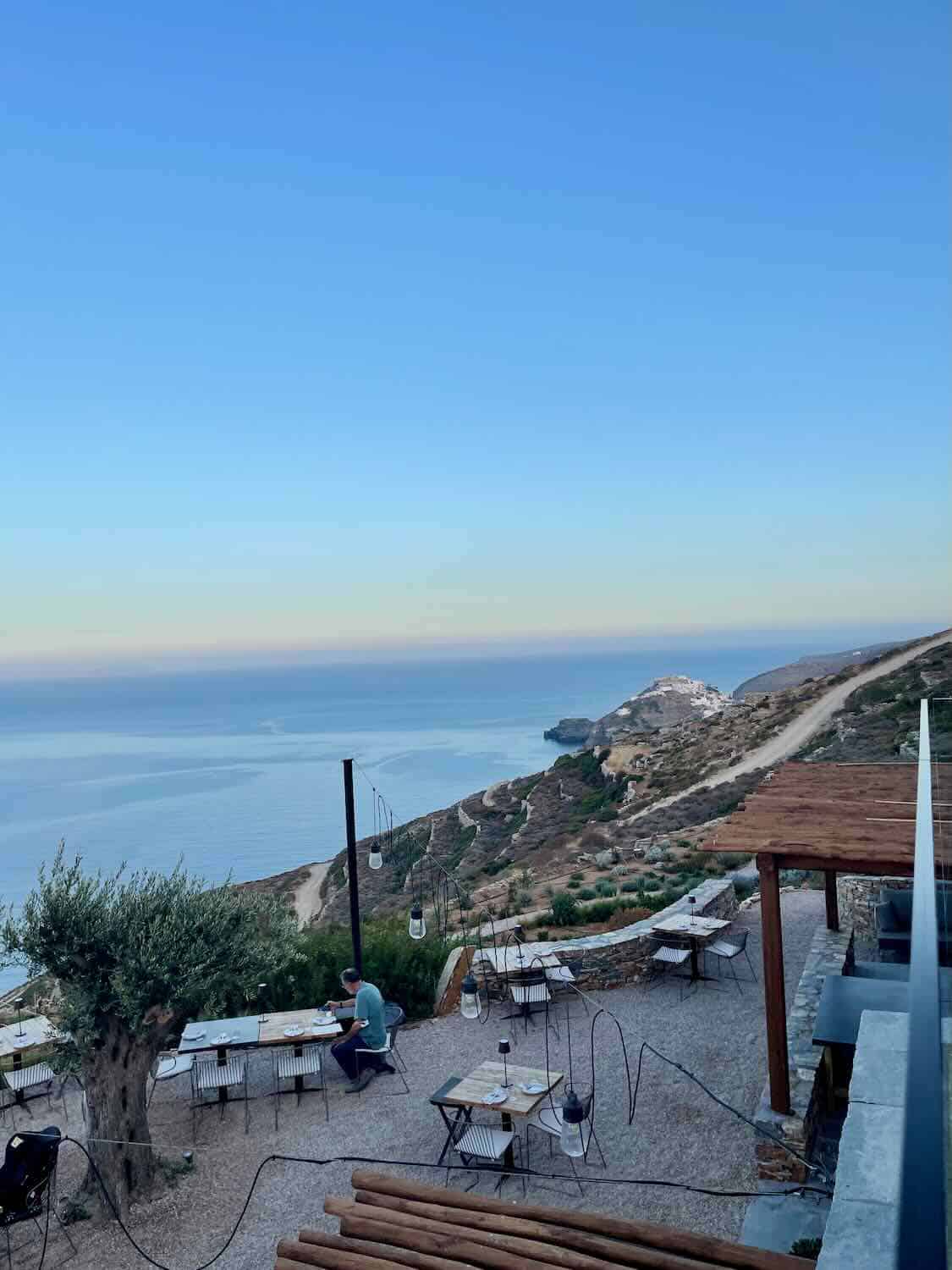 The image shows an outdoor dining area with tables and chairs arranged on a terrace overlooking a stunning coastal view. The scene features a calm, expansive sea under a clear blue sky, with a winding road visible along the cliffs in the distance. The seating area is shaded by wooden pergolas, creating a serene and inviting atmosphere, perfect for enjoying a meal or drink with a scenic backdrop.
