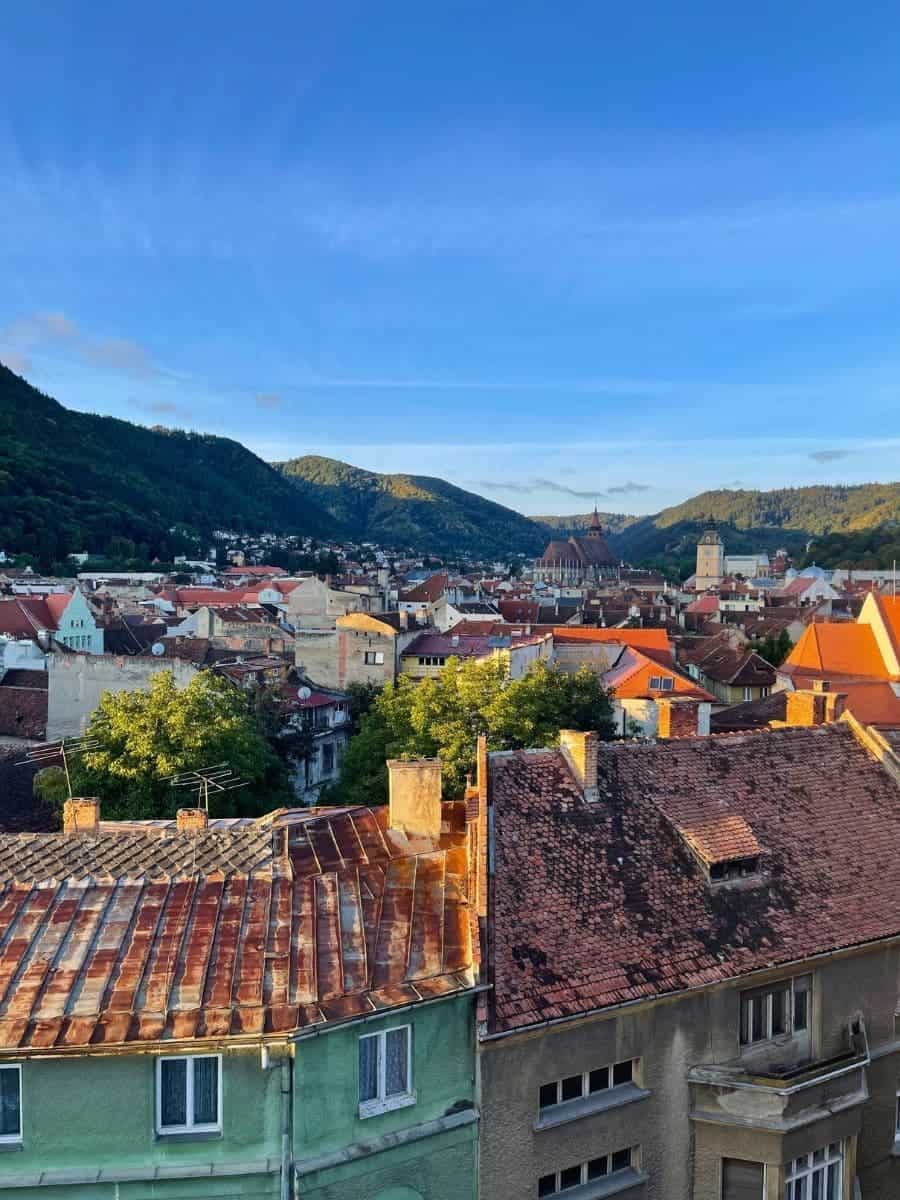 The image shows a scenic view of Bra?ov, Romania, in October, with the rooftops of traditional houses in the foreground. The rust-colored and weathered roofs complement the autumnal greenery of trees scattered among the buildings. In the background, rolling hills surround the town under a crisp, clear blue sky, capturing the beauty of the season.