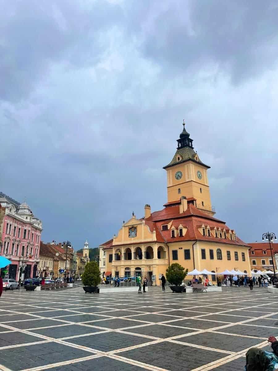 The image shows a central square in Brașov, Romania, featuring the historic Council House (Casa Sfatului) with its distinct clock tower and red-tiled roof. The square has a large open area with a grid-like pattern on the ground, surrounded by colorful, traditional buildings. The sky is overcast, giving the scene a moody yet striking atmosphere.