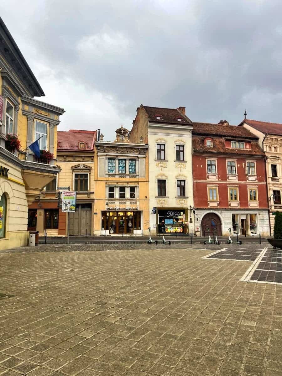 The image shows a quiet section of a square in Brașov, Romania, lined with colorful, historic buildings. The architecture is a mix of light pastel hues with charming, old-world facades. The street is paved with cobblestones, and the overcast sky adds a calm and serene atmosphere to the scene.
