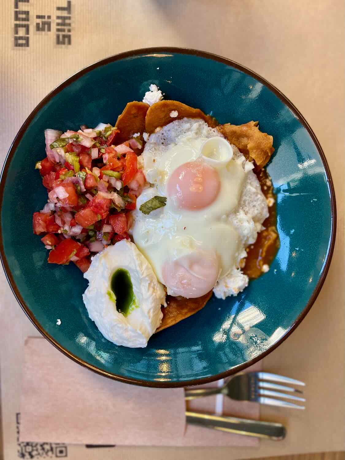 A close-up of a colorful dish featuring two poached eggs, fresh tomato salsa, and a dollop of yogurt with a drizzle of olive oil, served on a crispy base in a teal bowl.