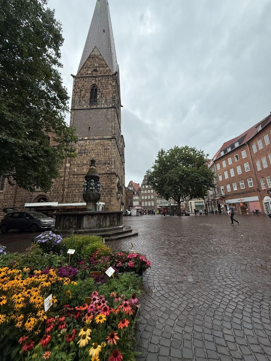 View of the stone church tower with a fountain in front, colorful flowers for sale, and the cobblestone square in Bremen.