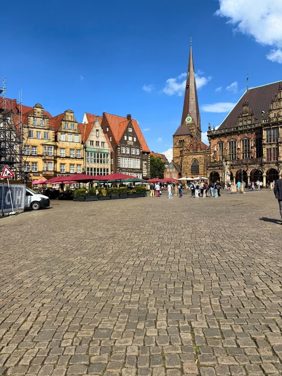 The bustling Bremen Market Square (Marktplatz) with its wide cobblestone plaza, colorful gabled houses, and the tall spire of St. Martin’s Church rising in the background. Crowds gather near the historic buildings and outdoor cafés.