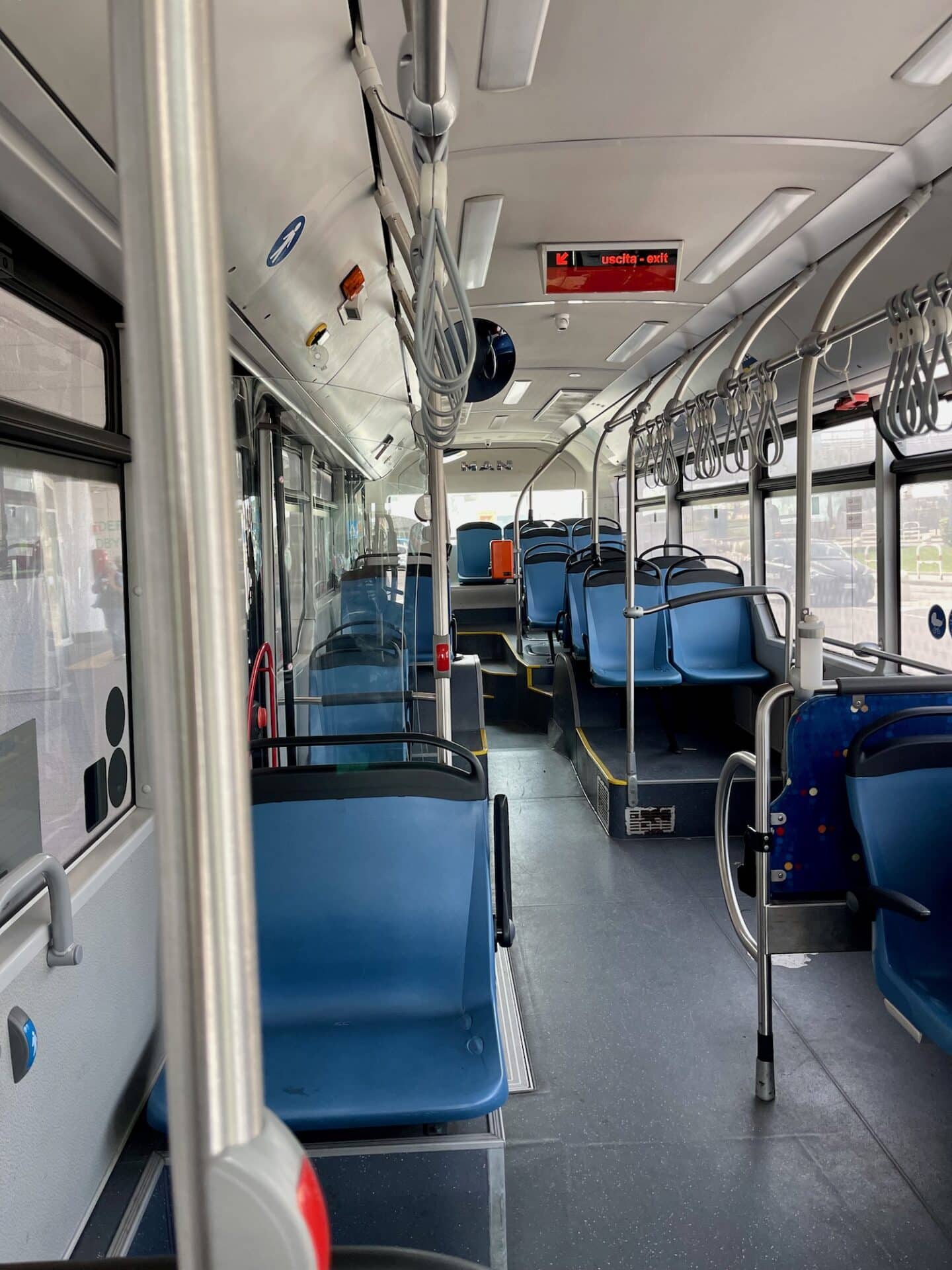 The interior of a modern city bus in Catania, Sicily, showing empty blue seats and metal handrails. The bus is well-lit, with large windows allowing sunlight to brighten the interior.