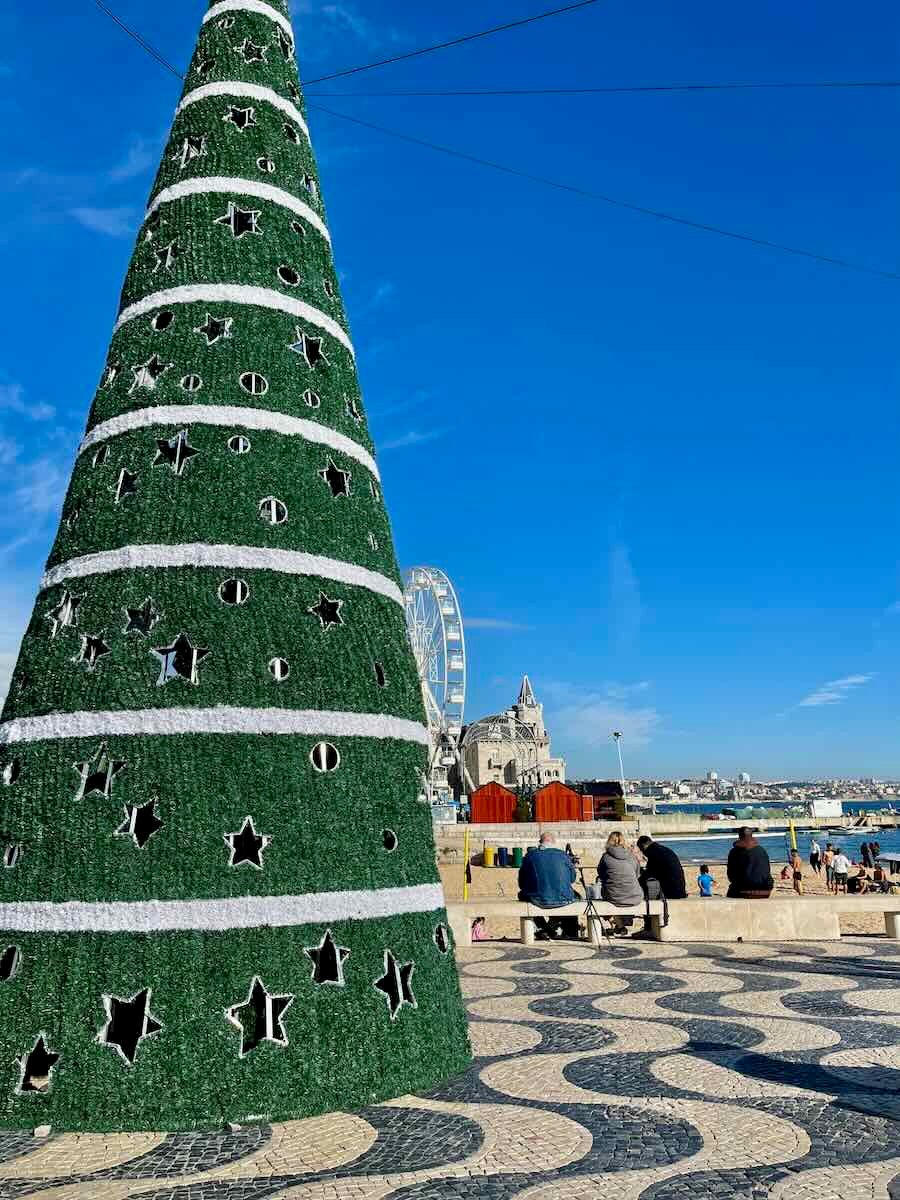 Large green Christmas tree with white accents standing near the Cascais waterfront, with a Ferris wheel and people sitting on a bench nearby.