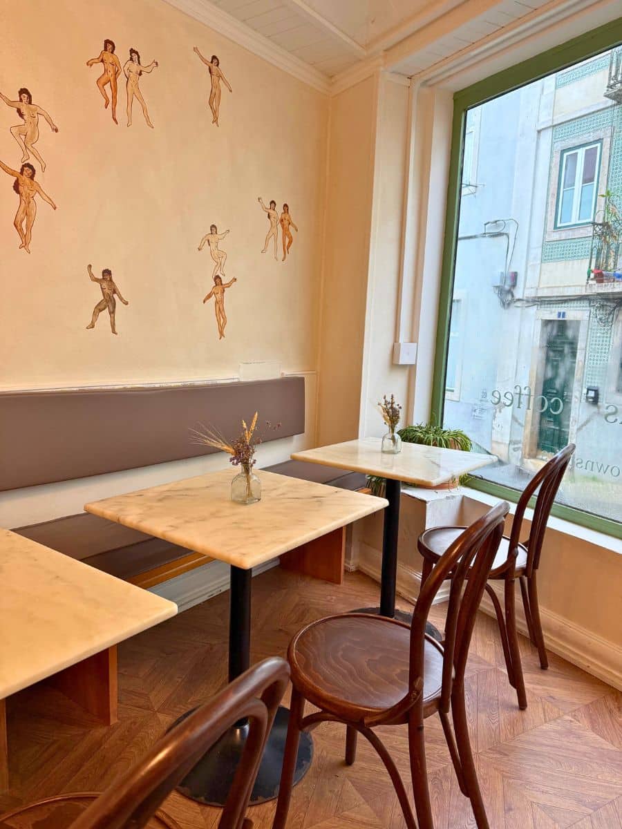 A cozy cafe in Lisbon with marble-top tables, wooden chairs, and a long cushioned bench. The wall features artistic illustrations of dancing figures, and a large window provides a view of the street outside. Small vases with dried flowers decorate the tables, creating a warm and inviting atmosphere.