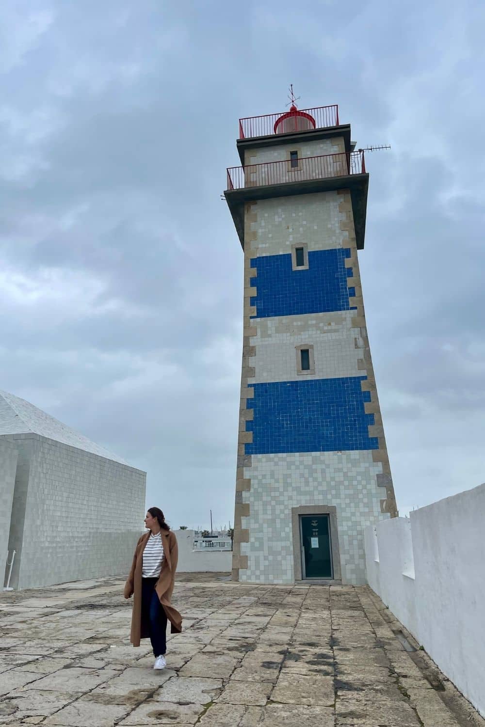 A person standing near the Santa Maria Lighthouse in Cascais, Portugal. The lighthouse features a stone exterior with blue and white horizontal stripes and a red light at the top. The sky is overcast, adding to the quiet and serene atmosphere.