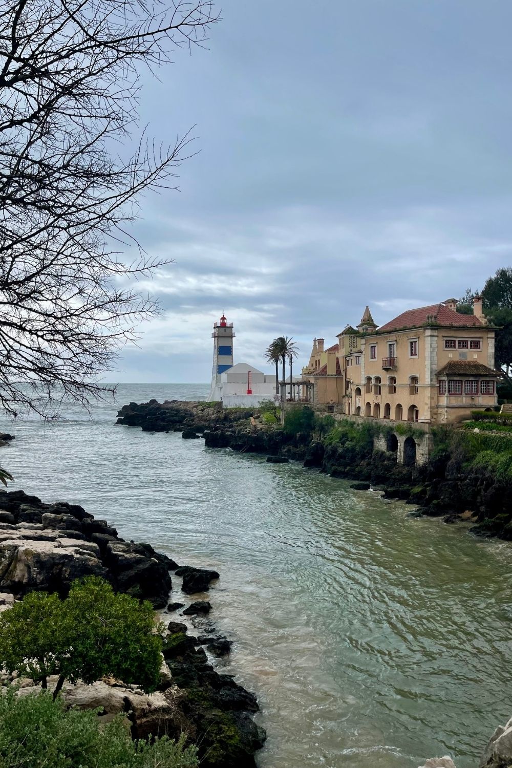 A scenic view of the Santa Maria Lighthouse on a rocky coastline in Cascais, Portugal, with the ocean on one side and historic buildings, including a villa, on the other. The sky is cloudy, and the water is calm, creating a tranquil atmosphere.