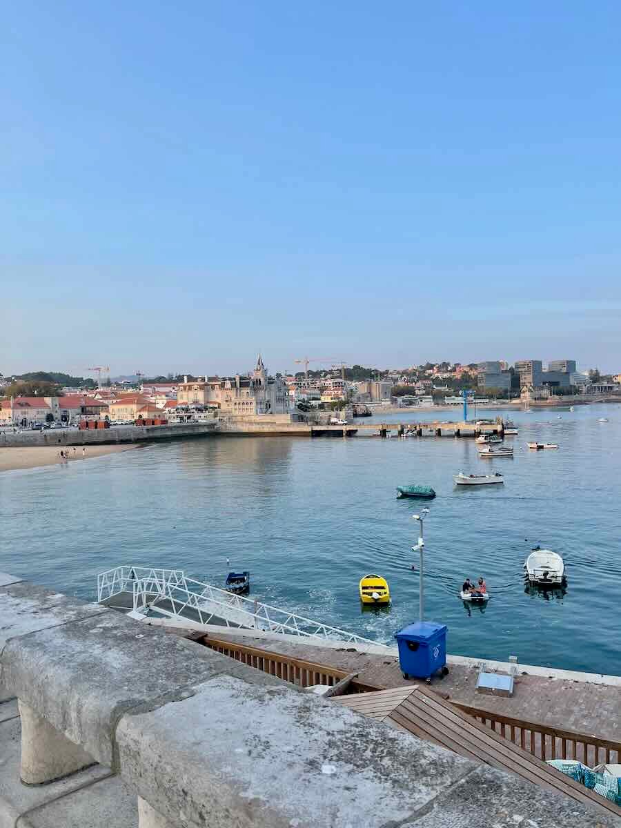 A serene view of the waterfront in Cascais, Portugal. Small boats are floating peacefully in the calm harbor, with the town's buildings visible in the background, under a pastel sky as the evening sets in.