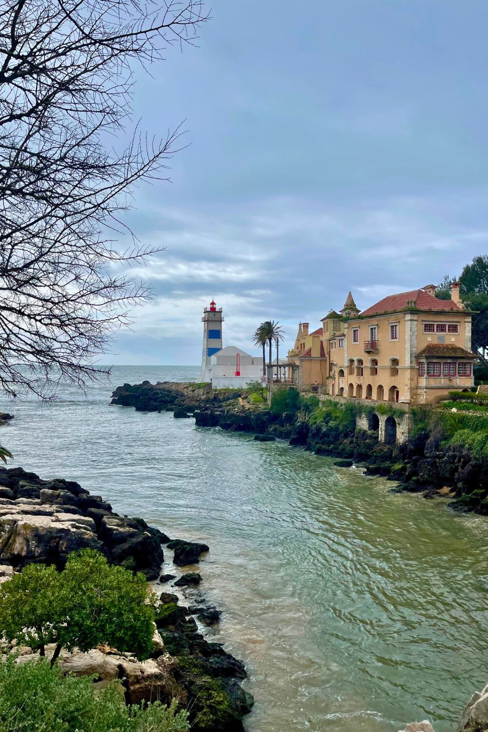 A scenic coastal view in Cascais, Portugal, with a lighthouse, historic buildings along the water, and cloudy skies, creating a tranquil atmosphere.