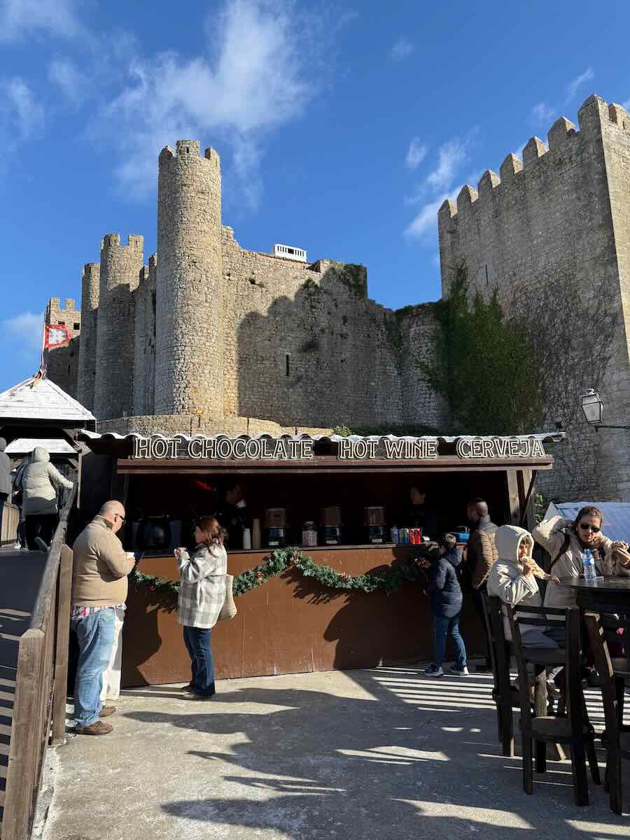A cozy Christmas market stall selling hot chocolate, mulled wine, and beer, set against the backdrop of historic castle walls under a bright blue sky.
