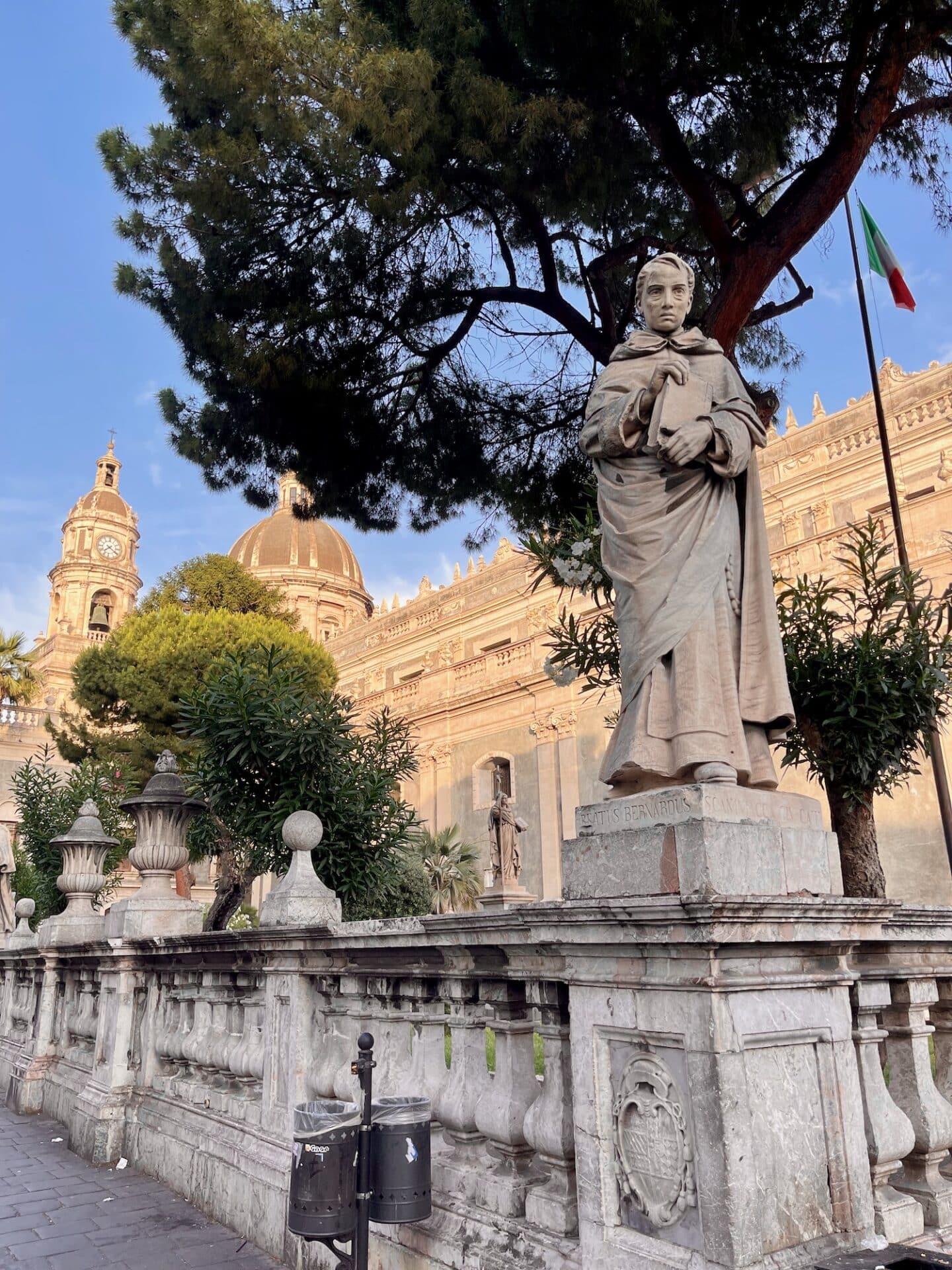 Statue in the gardens near Catania Cathedral, surrounded by lush trees and historic architecture, with the Italian flag subtly visible in the background under a soft afternoon light.