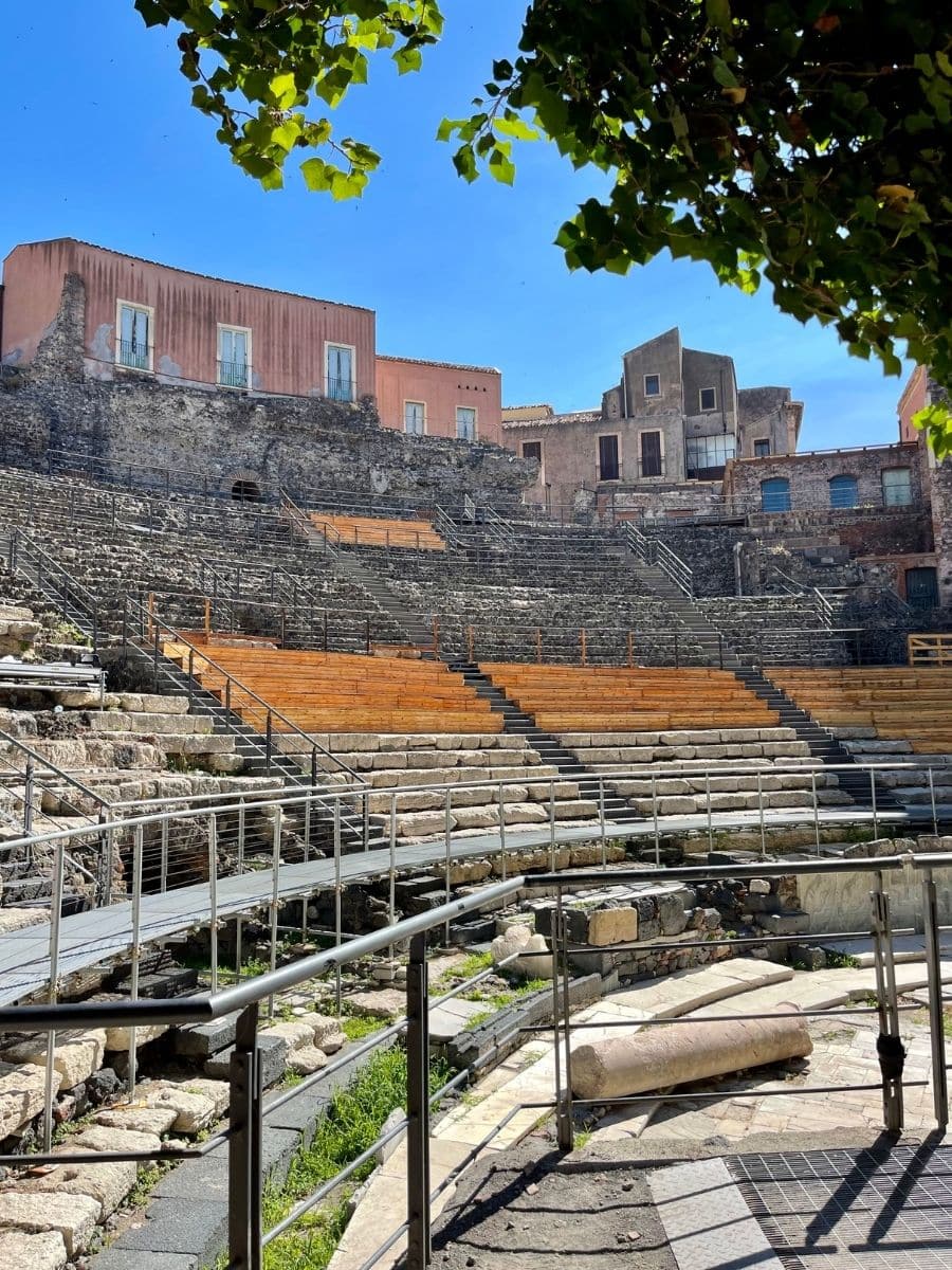 Ancient Roman amphitheater in Catania, Italy, with stone seating, modern railings, and historic buildings in the background under a bright blue sky.