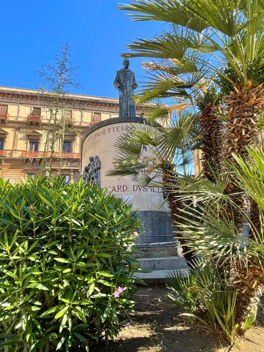 Statue of Cardinal Dusmet in Catania, Italy, surrounded by lush green plants and palm trees, with a historic building in the background under a clear blue sky.