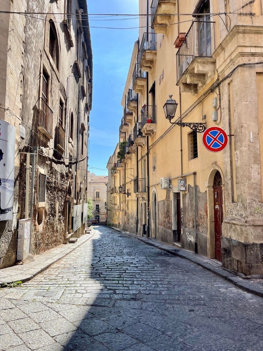 A narrow cobblestone street in Catania, Sicily, lined with historic buildings showcasing a mix of weathered and well-preserved facades under a bright blue sky.