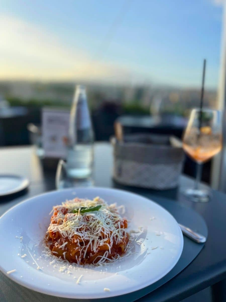 A plate of pasta topped with tomato sauce, grated cheese, and fresh basil, served at an outdoor restaurant in Catania with a scenic sunset view in the background.