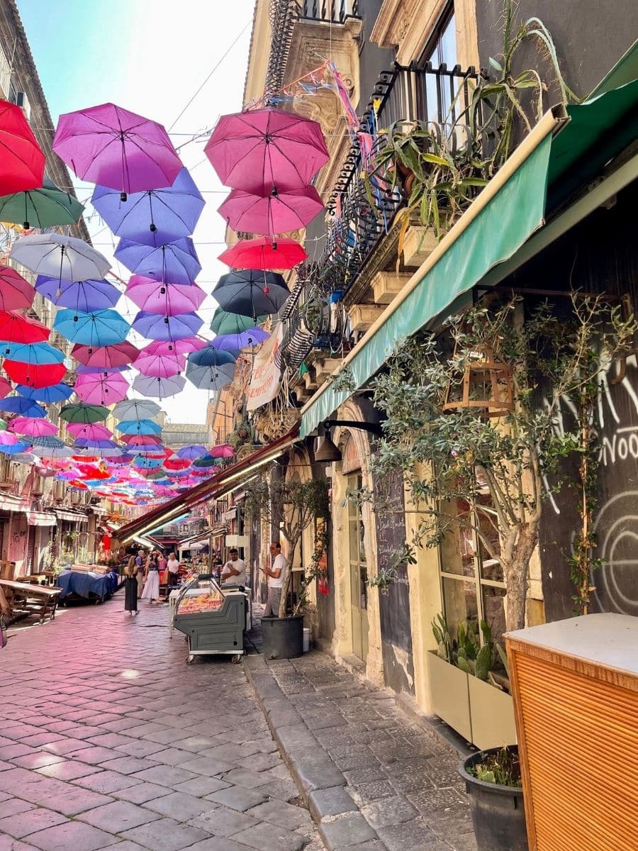 Vibrant street in Catania, Sicily, decorated with colorful umbrellas hanging overhead, lined with charming cafés, market stalls, and historic architecture.