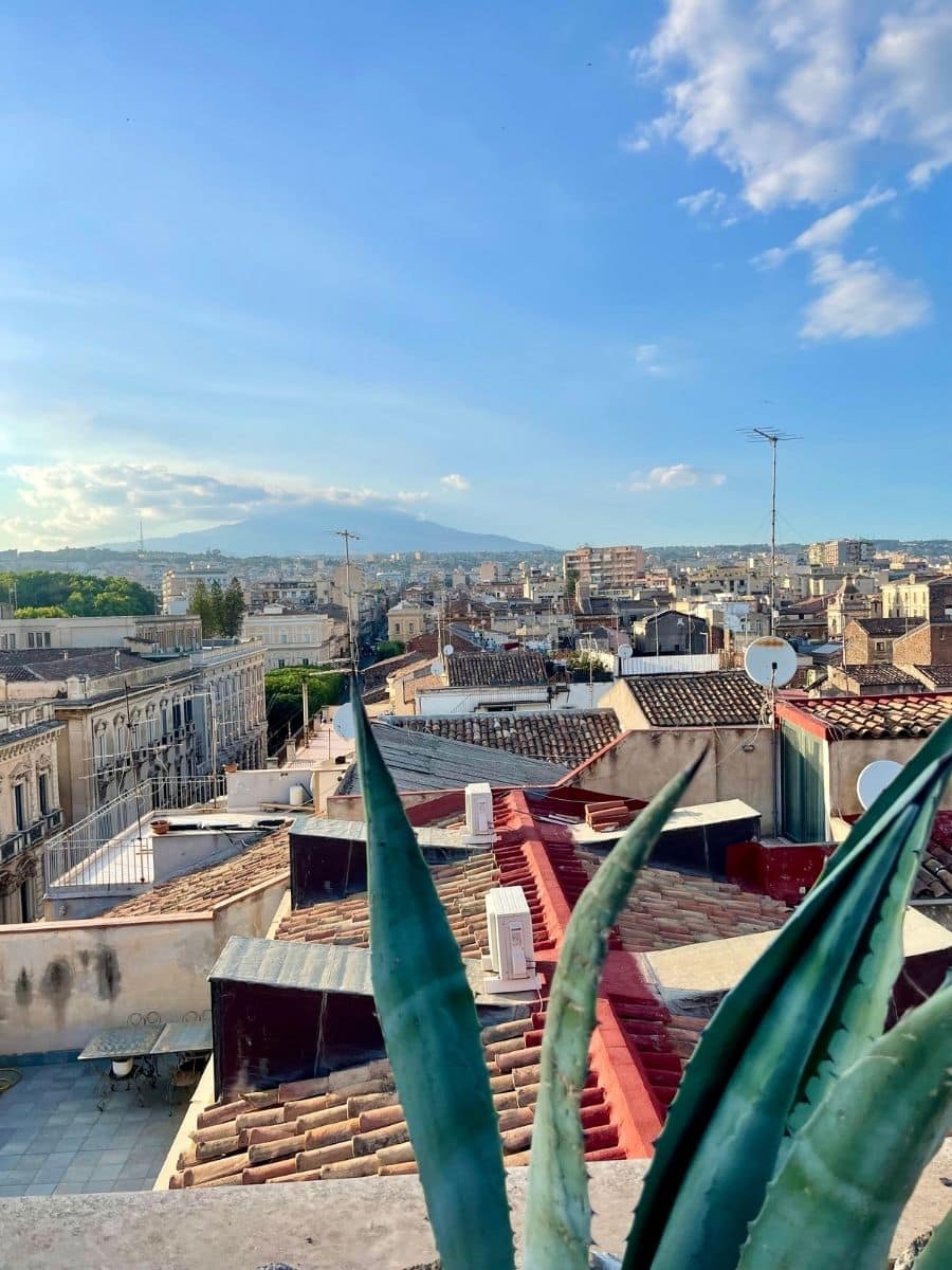 Scenic rooftop view of Catania, Sicily, with historic buildings, terracotta rooftops, and Mount Etna in the background under a clear blue sky.