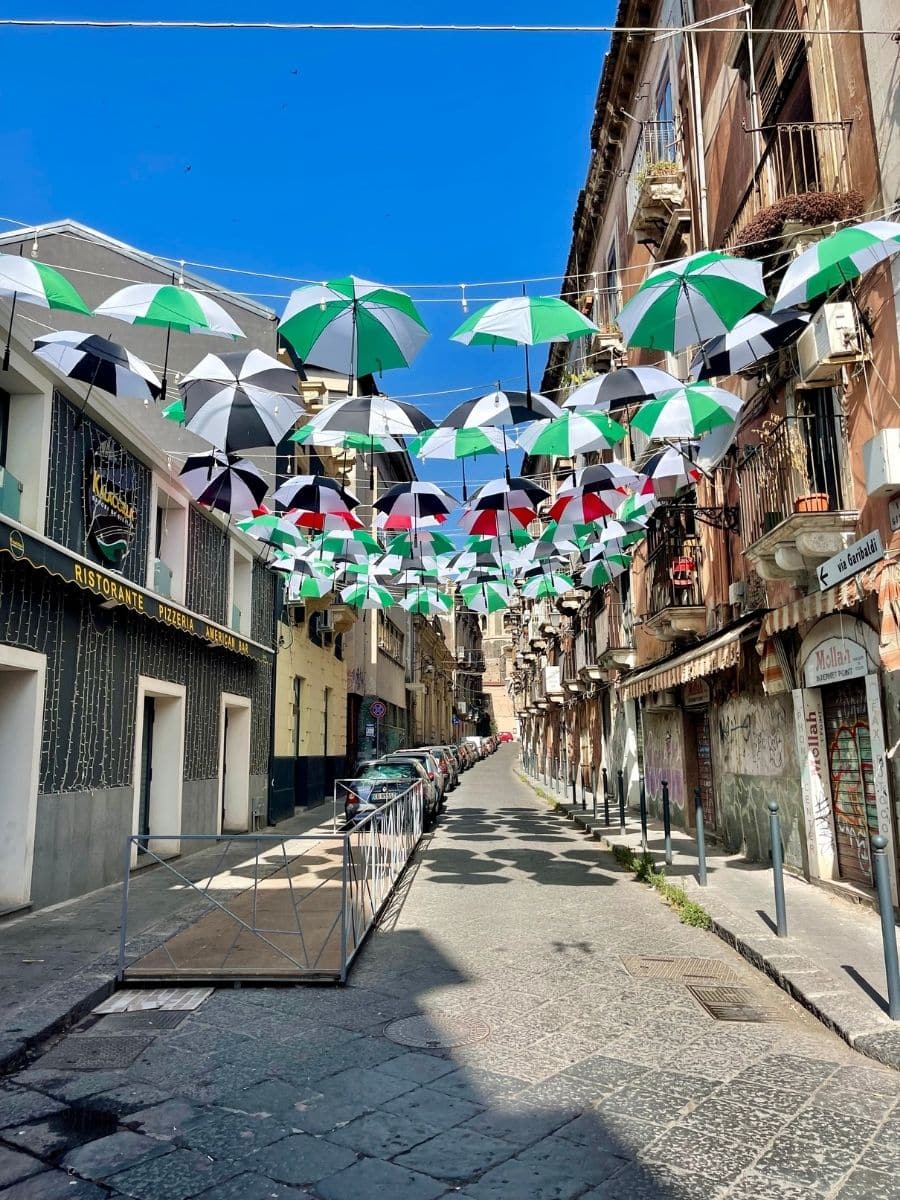 Street scene in Catania, Sicily, featuring a "Ristorante Pizzeria" sign on the left and a decorative display of hanging umbrellas in various colors.