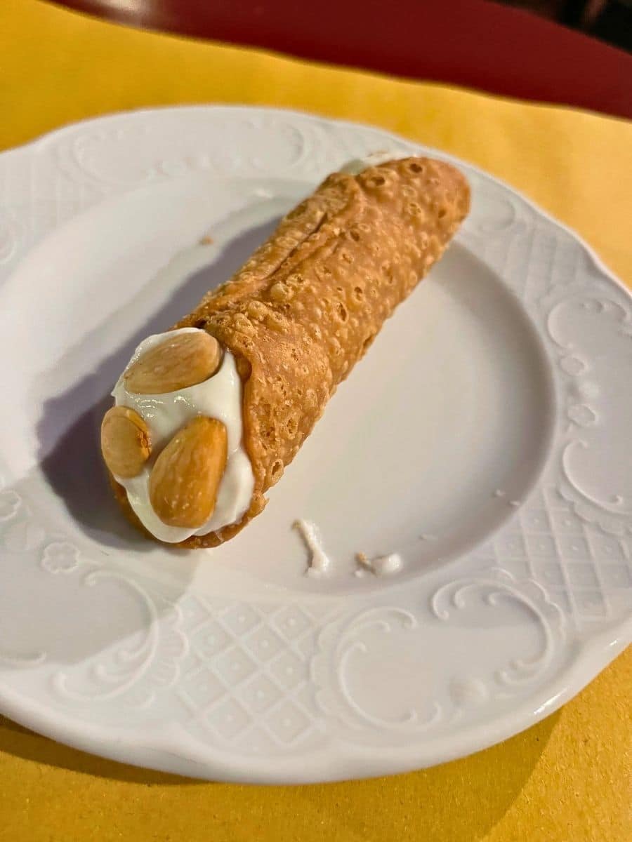 Close-up shot of a crispy cannolo shell filled with creamy ricotta cheese and garnished with almonds, served on a decorative plate in Catania, Sicily.