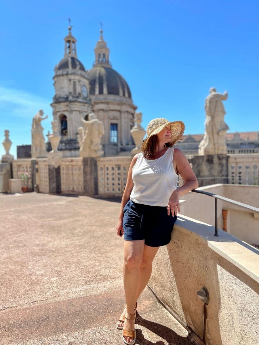 Melissa wearing a straw hat, white sleeveless top, and navy shorts leans against a stone railing on a sunny terrace in Catania, Sicily. Behind her, the grand domes and statues of an ornate historic church stand against a clear blue sky, highlighting the city's rich Baroque architecture.