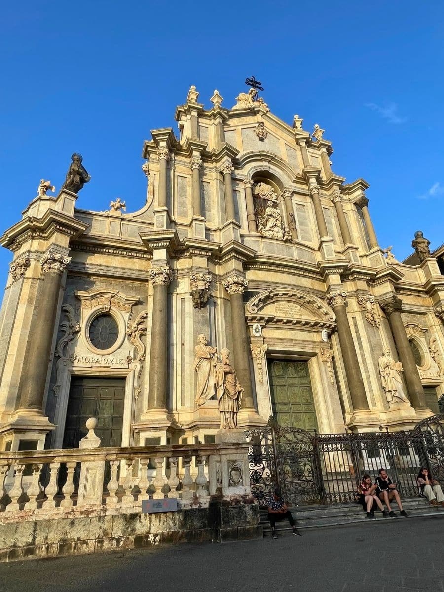 A view of the Catania Cathedral in Piazza del Duomo, Catania, Sicily, showcasing its baroque architecture, statues, the "ALOPA OVLE" inscription, and the surrounding piazza with its paving stones and people sitting on the steps.